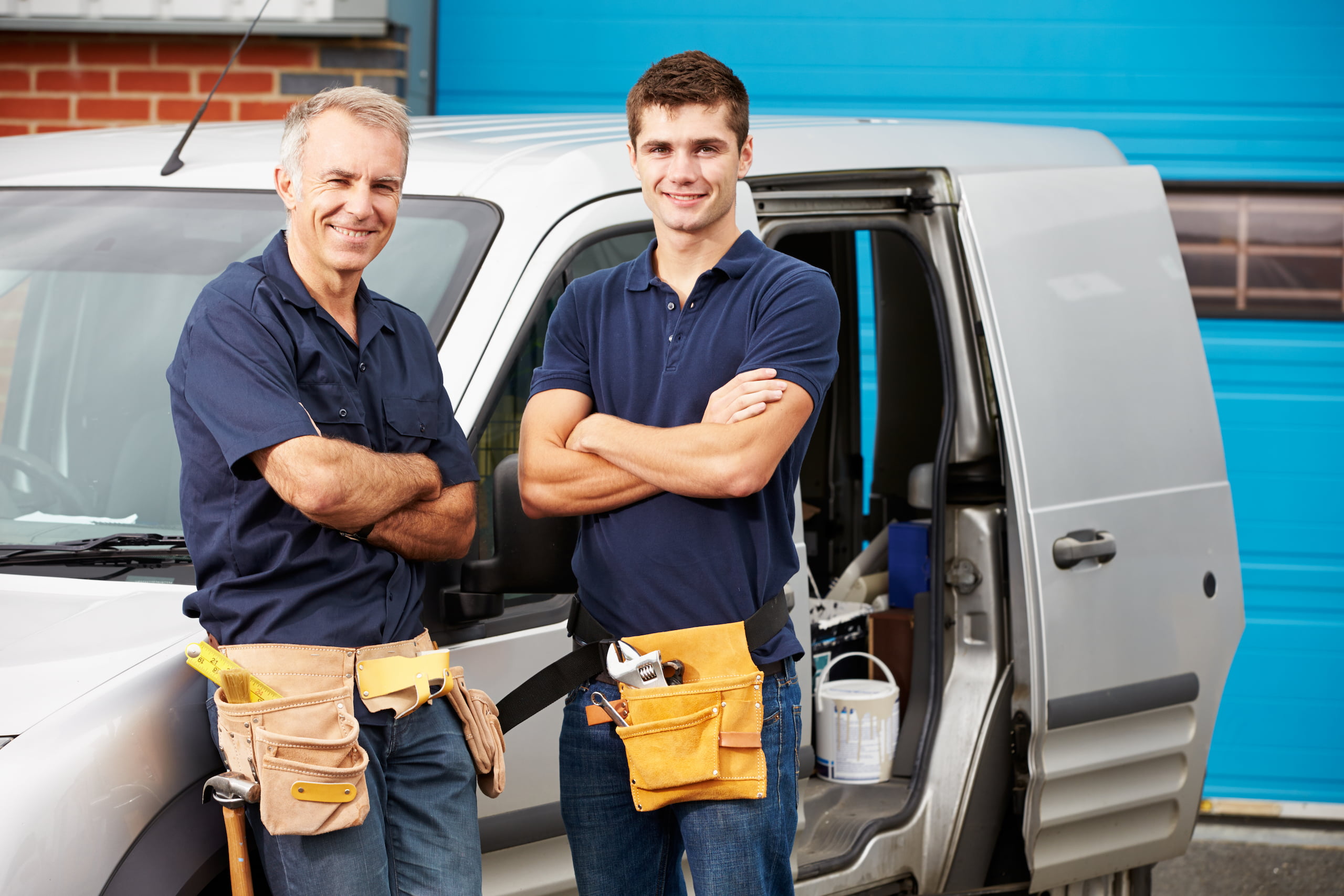 Two men proudly stand smiling in front of a van with its door open, their work uniforms and tool belts signaling their role as dedicated workers in a family business.