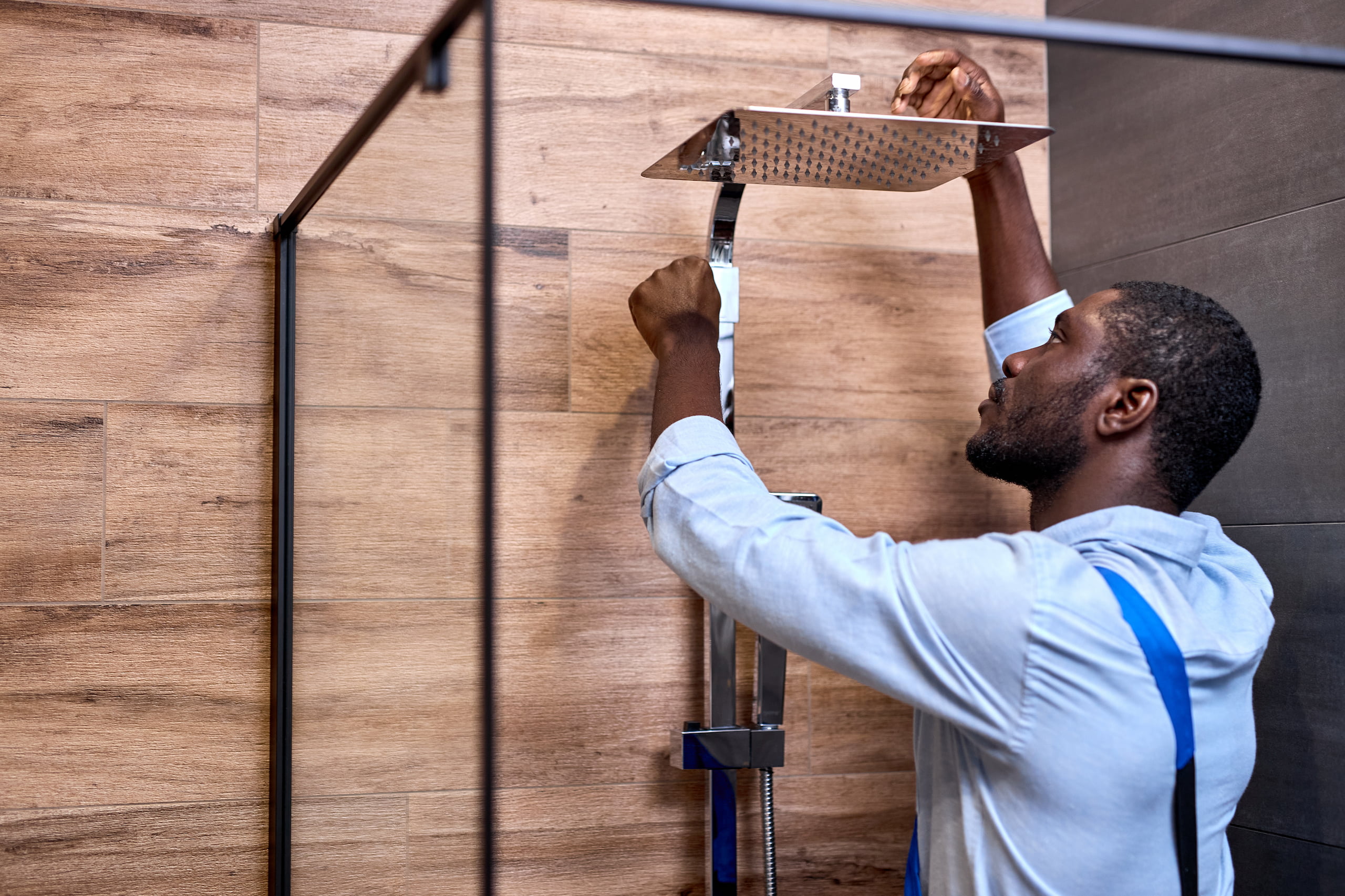 A plumber in a blue shirt is skillfully installing a square showerhead in a modern bathroom adorned with elegant wood paneling.