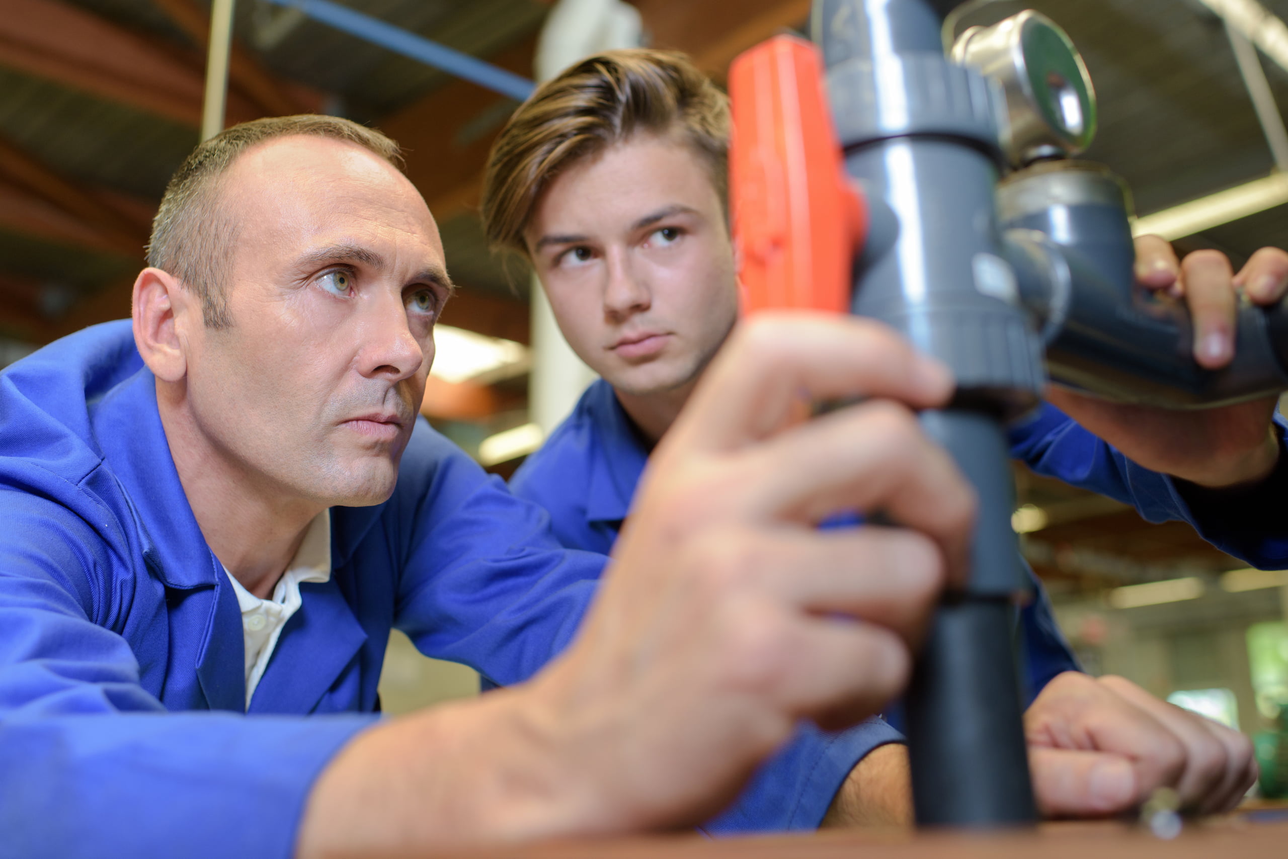 Two men in blue coveralls, resembling a professor and student in a craft class, are closely inspecting a plumbing pipe inside the workshop. One man gestures towards the pipe with focused attention.