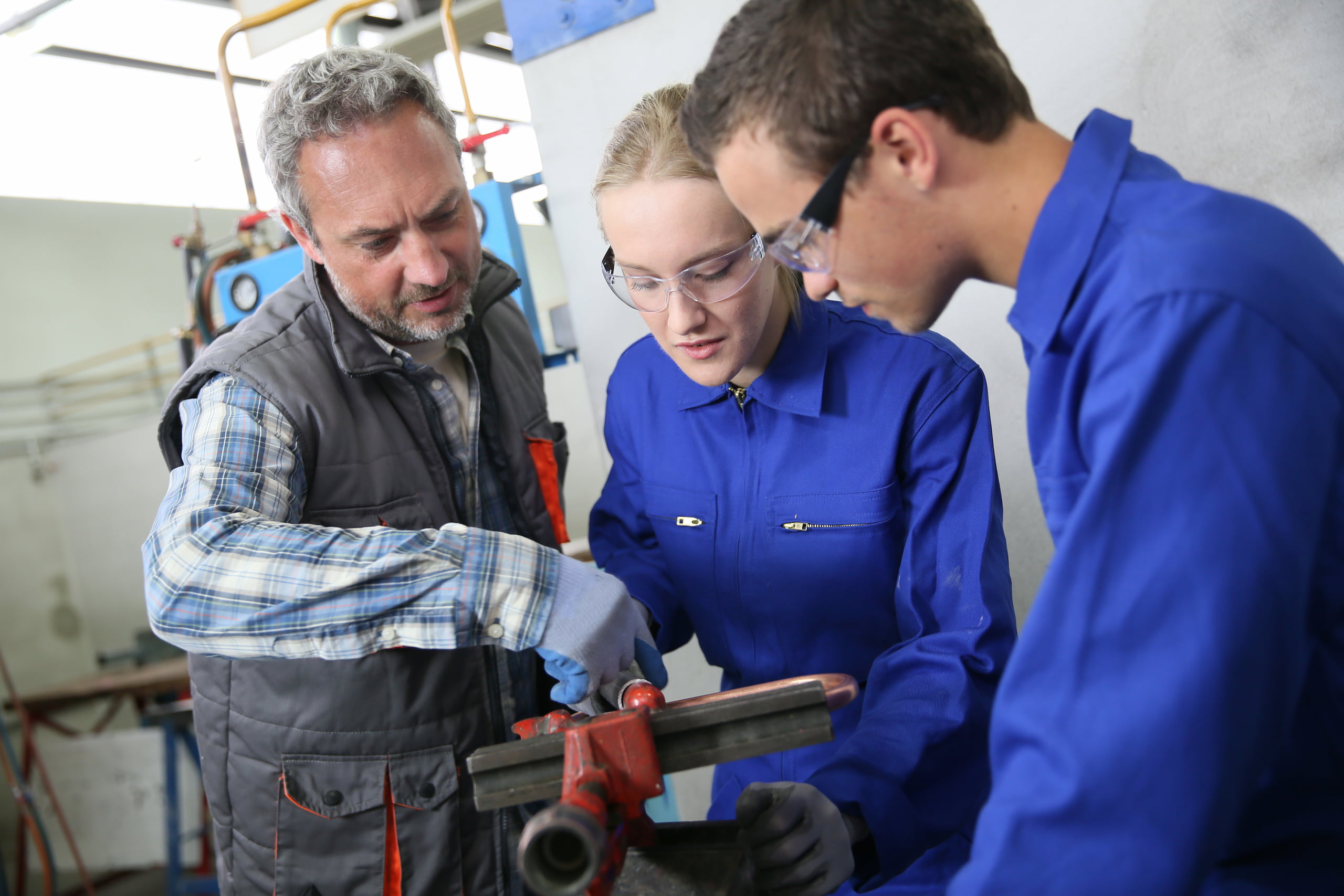 An instructor guides two students in blue overalls working with a vise in a workshop, as the students in plumbing focus intently on listening to the teacher's instructions.