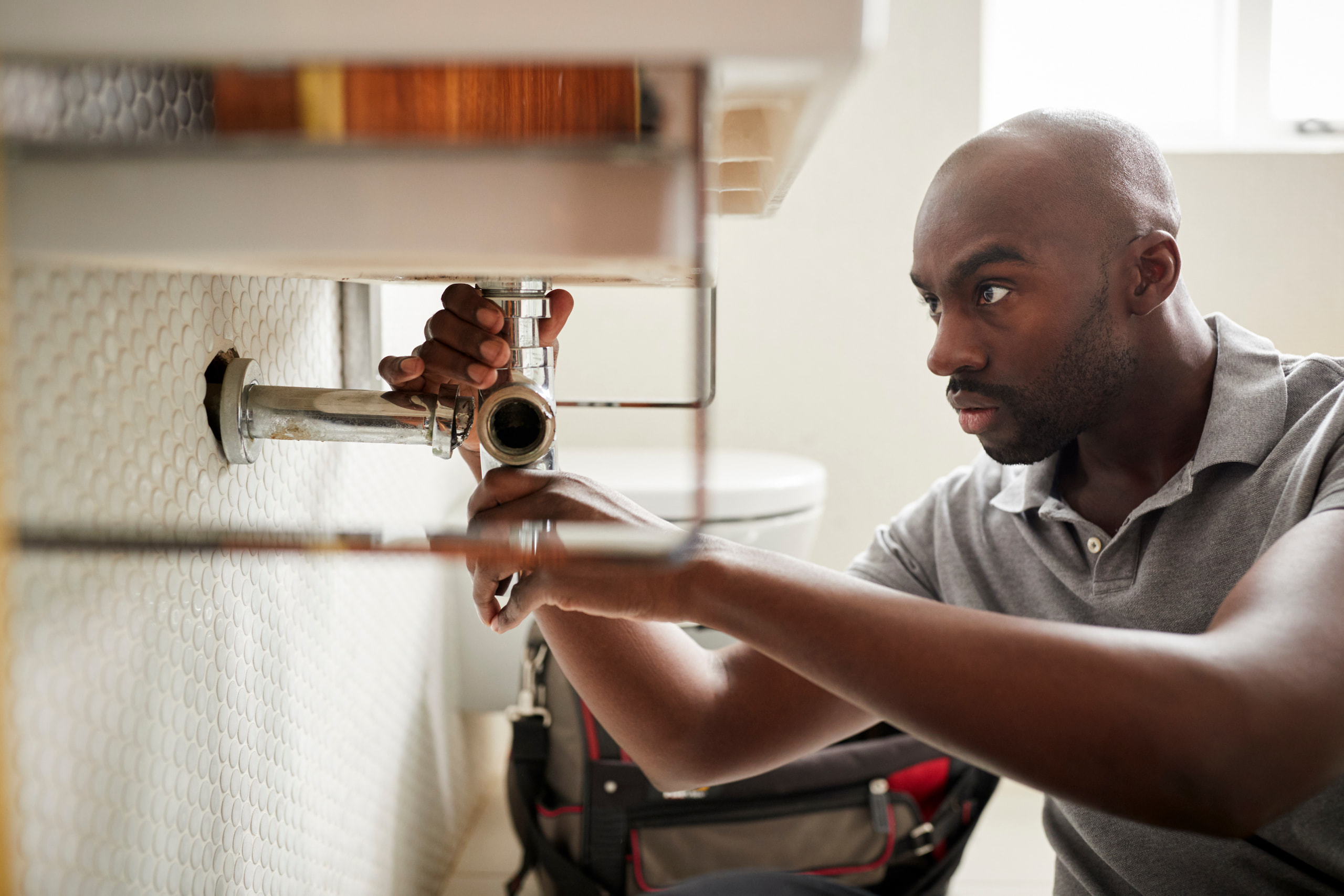 A man in a gray shirt fixes a pipe under a sink with focused attention. A toolbox is visible in the background.