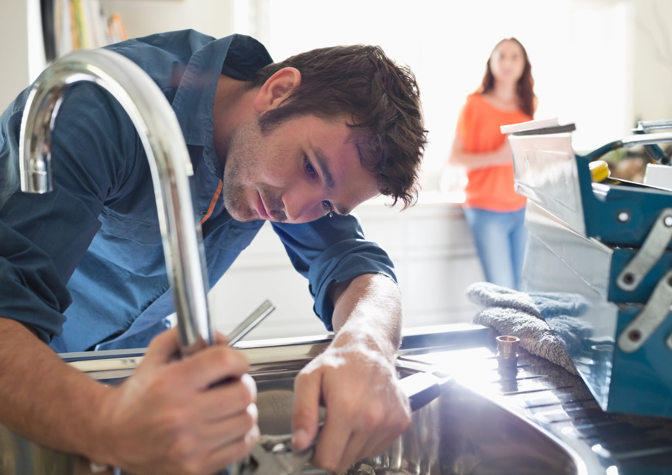 A man in a blue shirt repairs a kitchen sink, focused on using wrenches. A woman in an orange top stands in the background, watching. A toolbox is nearby on the counter.
