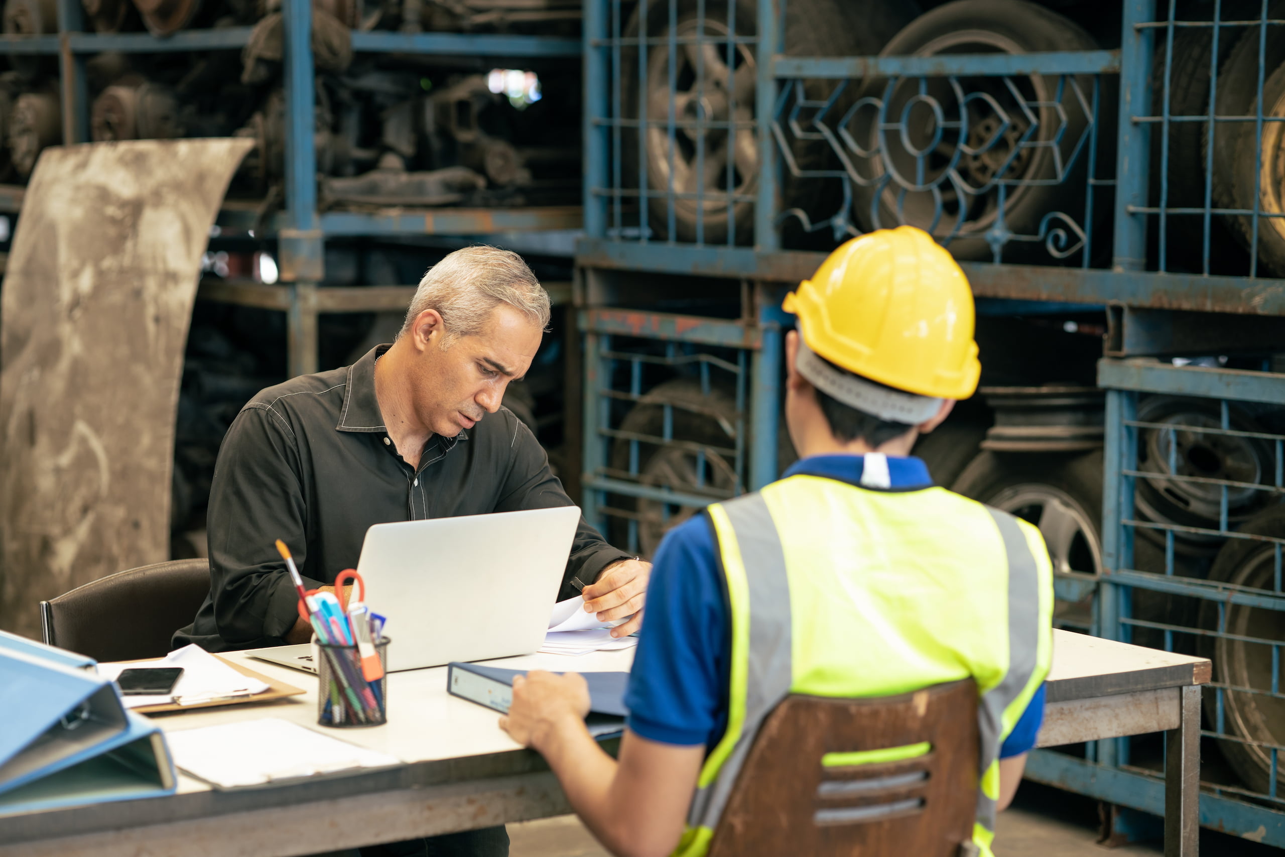 A man works on a laptop at a desk, facing a person in a hard hat and safety vest. Amidst shelves holding machine parts, their discussion subtly incorporates interview coaching tips, bridging industrial knowledge with essential conversational skills.