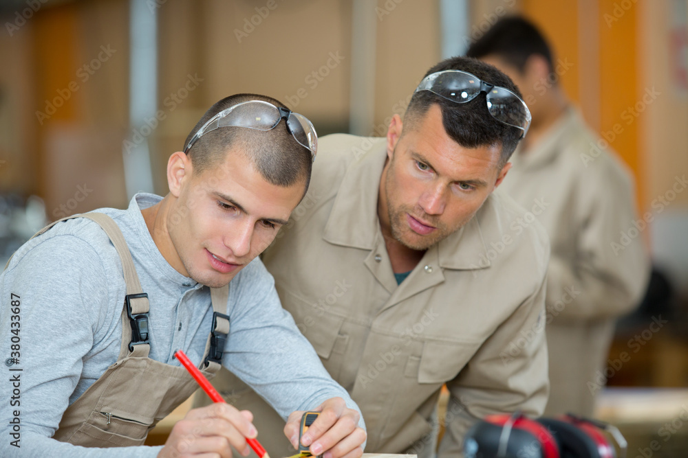 Two men in work clothes and safety goggles collaborate on a woodworking project. One draws on wood with a pencil while the other observes.