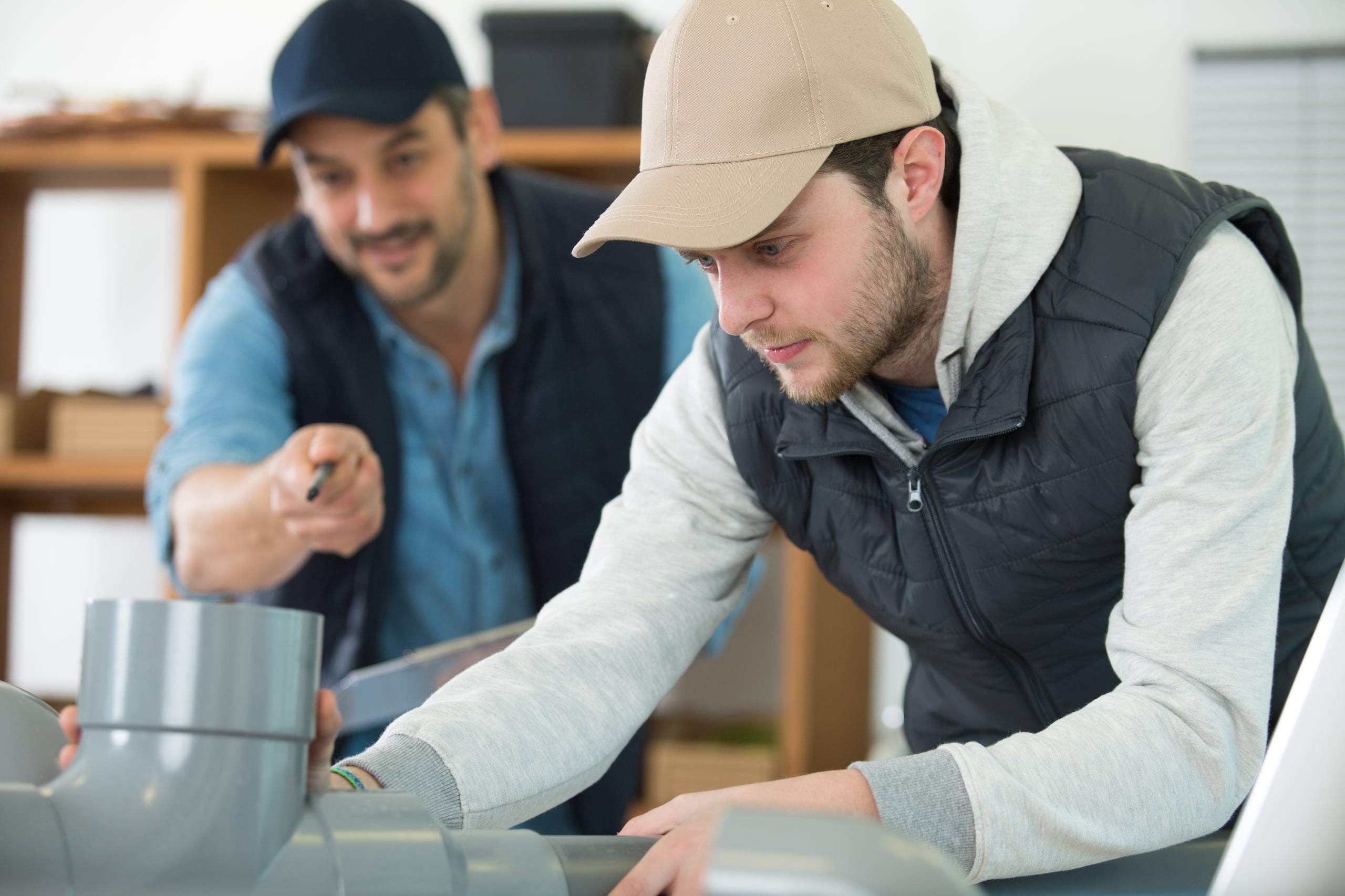 Two men working together on a plumbing project; one points at a pipe section while the other adjusts it. Both wear caps and casual work attire.