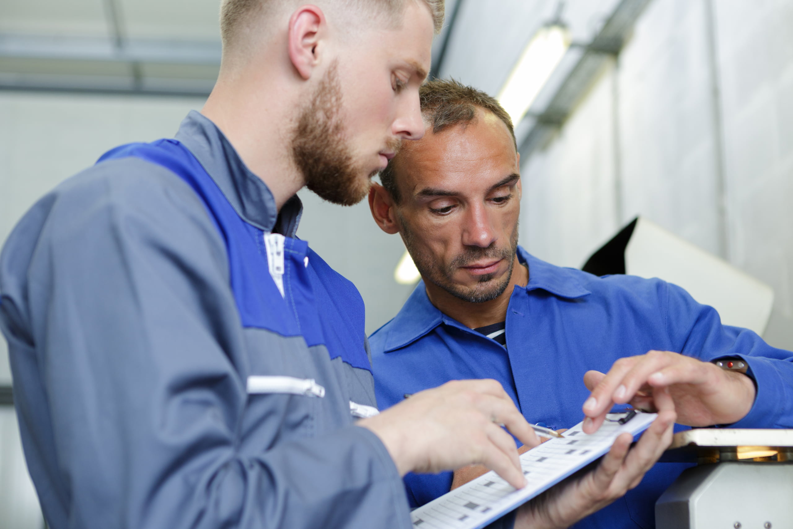 Two men in plaid shirts, one holding a wrench and the other holding a faucet, discuss plumbing repairs in a white-tiled room.