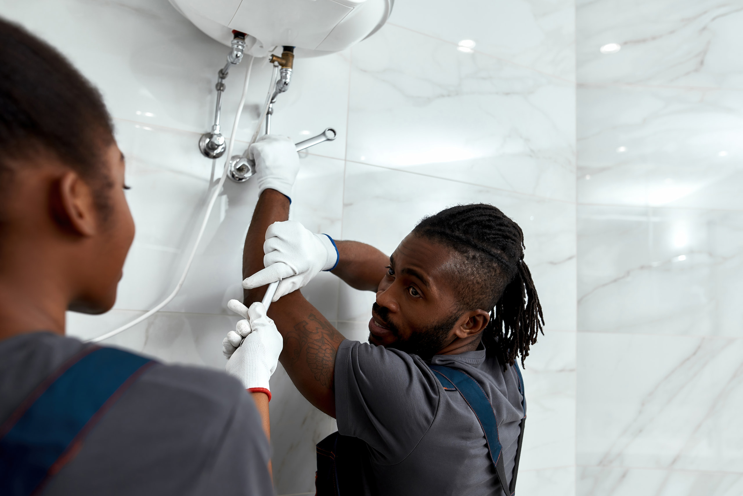 Two plumbers in uniforms and gloves fixing pipes under a sink in a tiled bathroom. One is adjusting the pipes while the other observes.