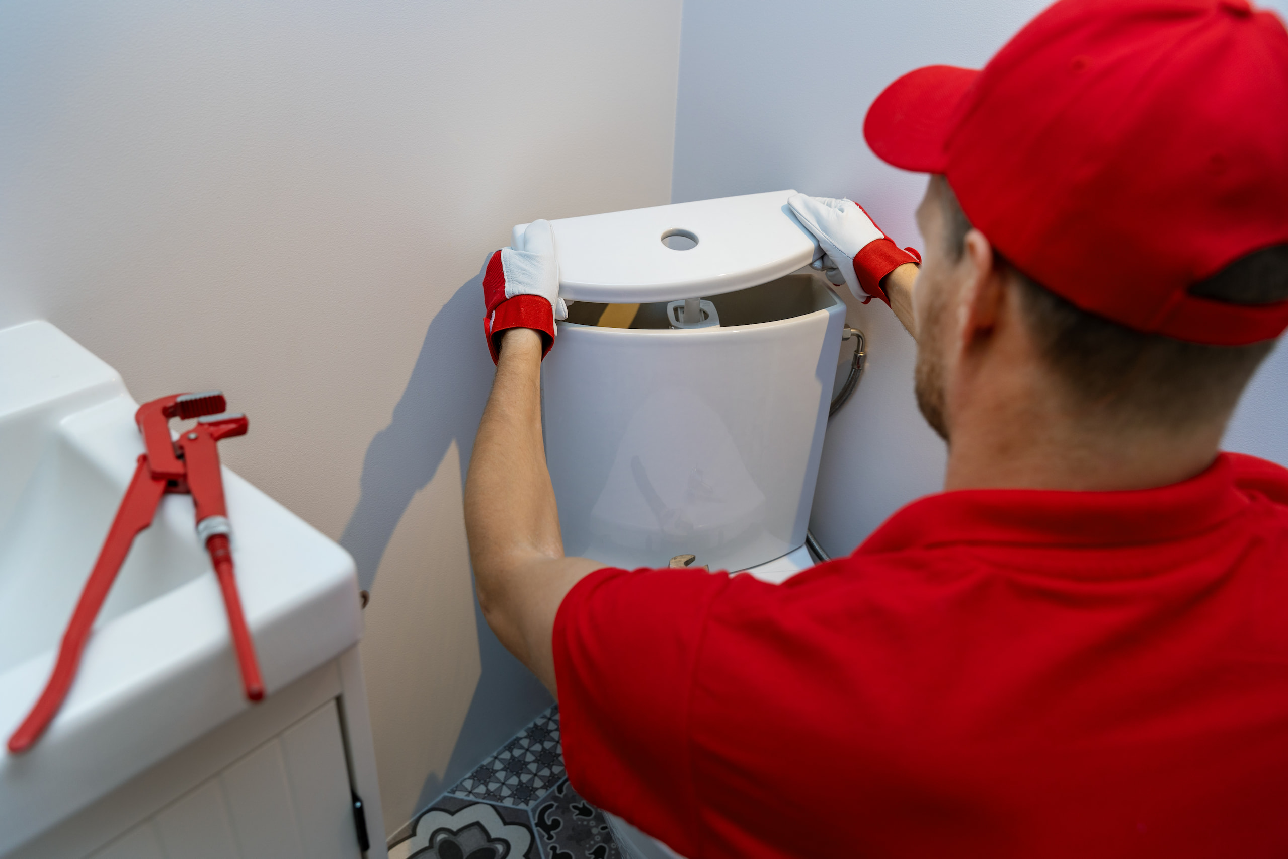 Worker in red uniform fixing a toilet tank in a bathroom, with plumbing tools nearby.