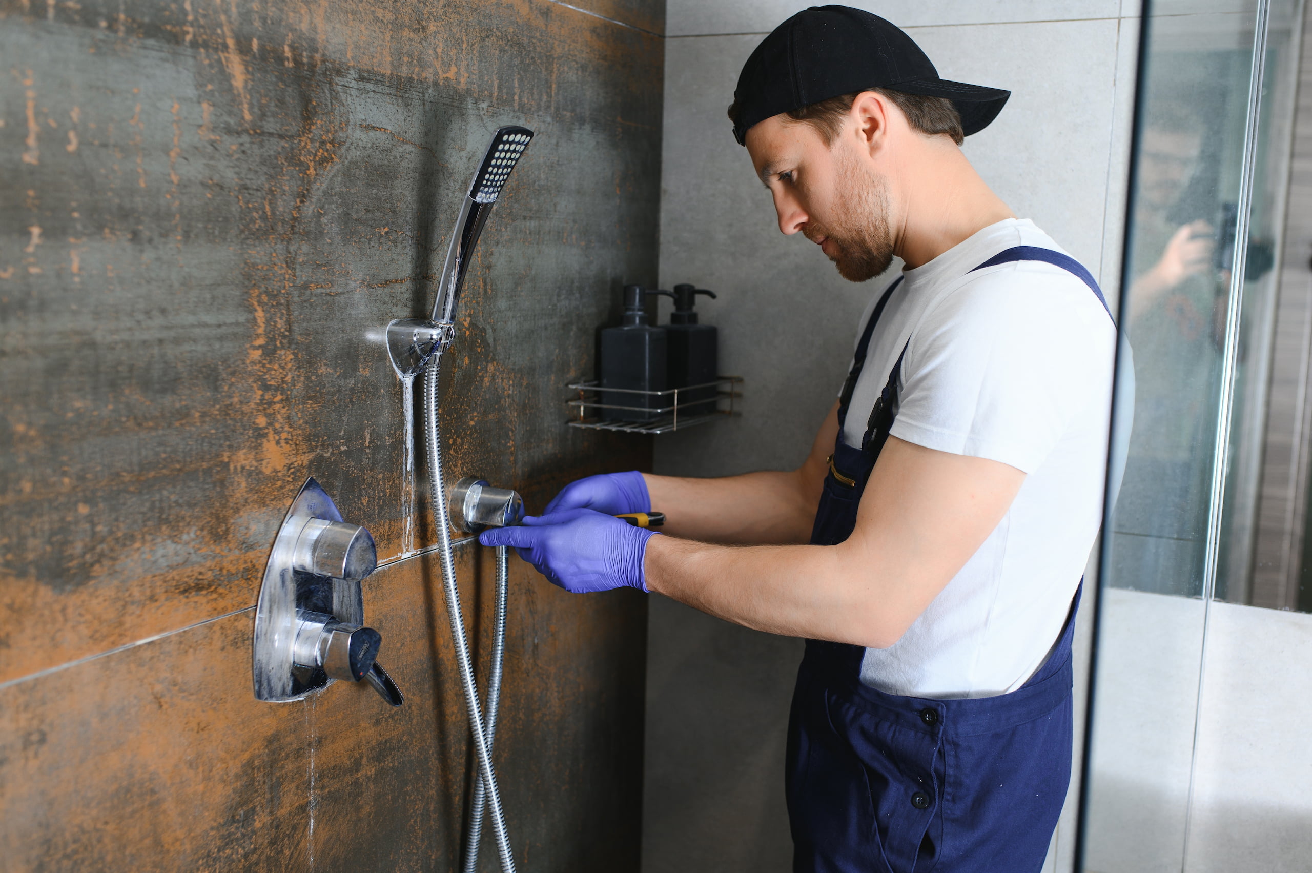 Man in overalls and gloves repairs a shower in a bathroom.