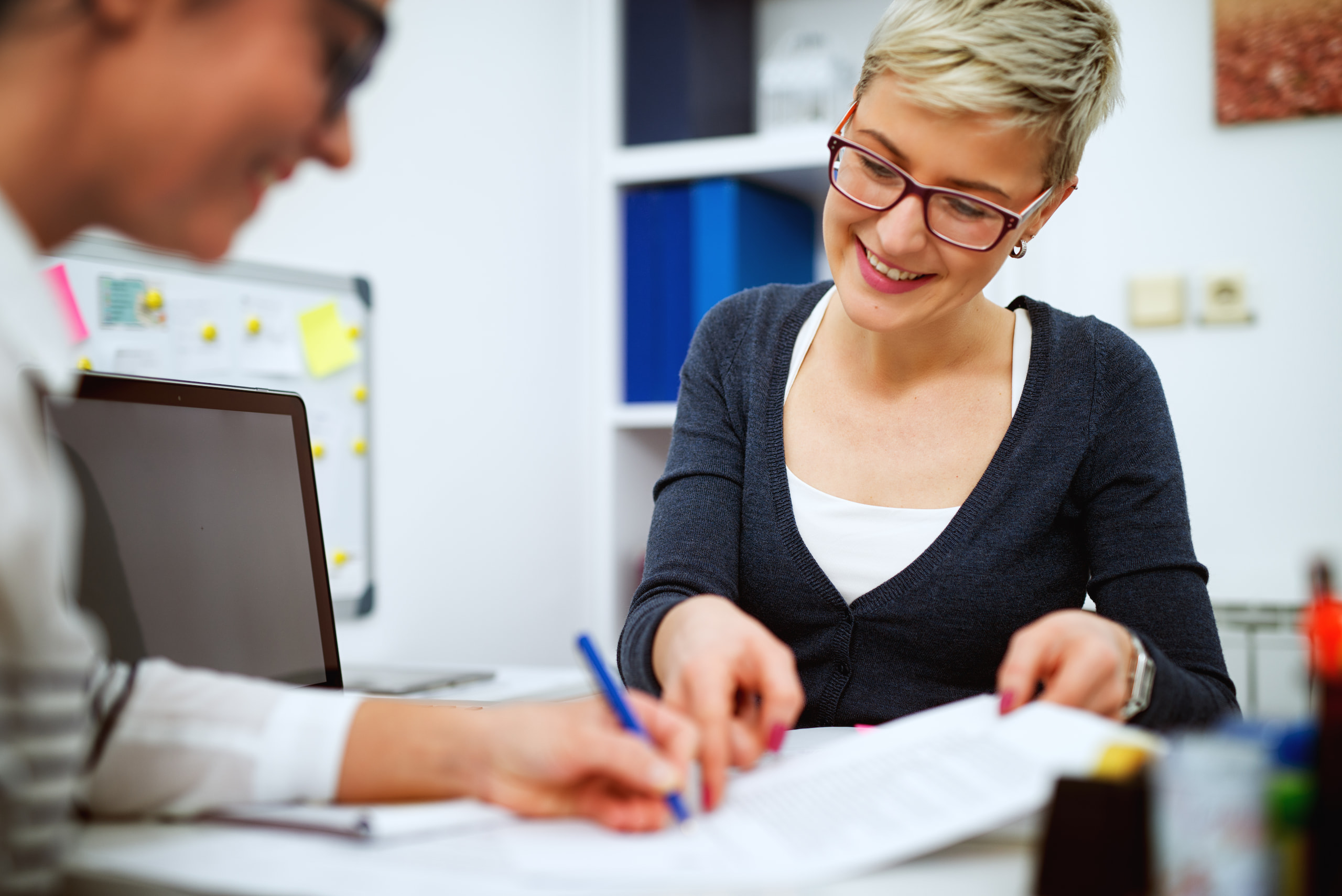 Two women at a desk in an office setting are getting started on their project, discussing papers. One woman is smiling and pointing, while the other is writing. A laptop and office supplies are visible in the background.