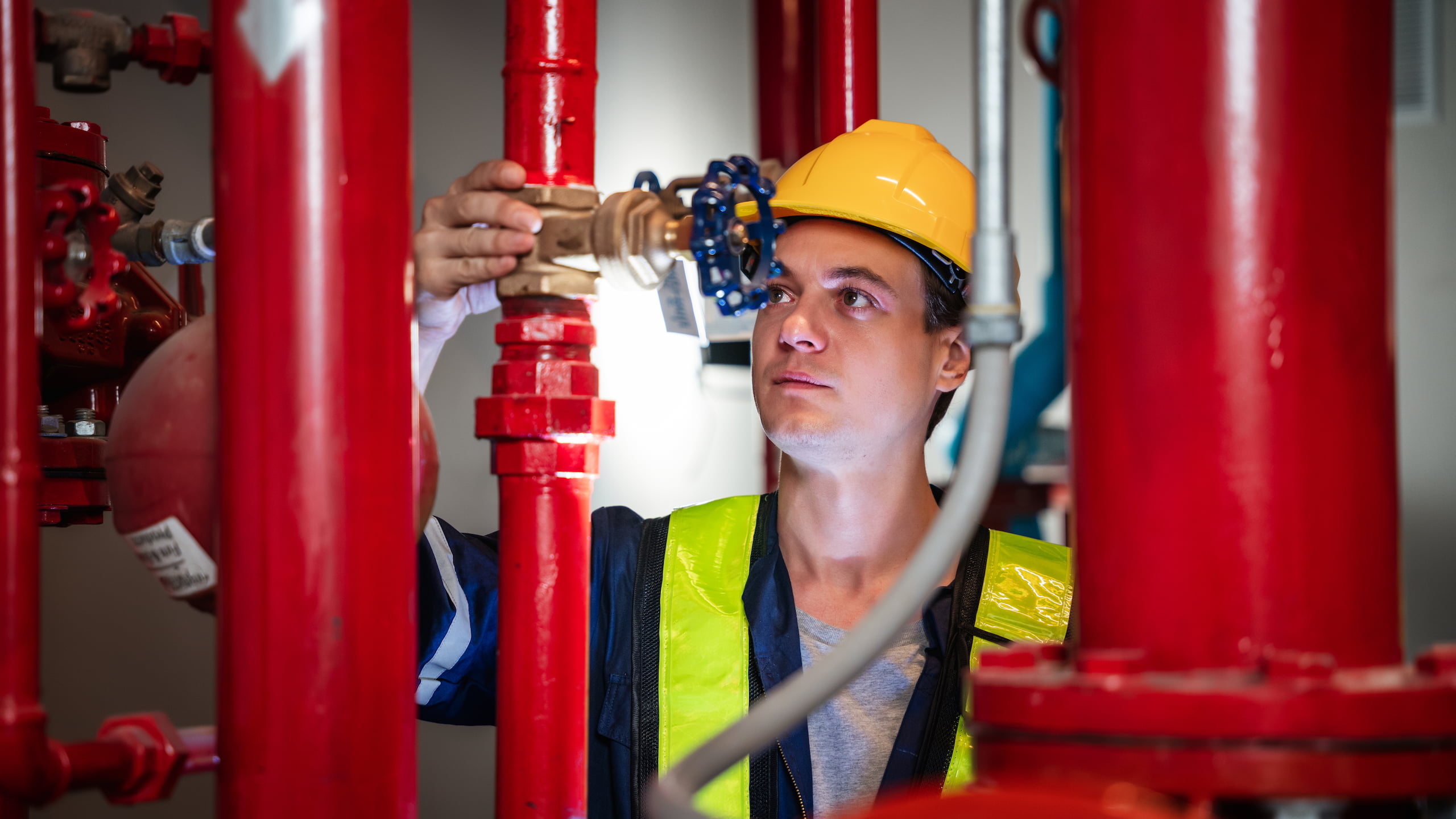 A technician in a hard hat and safety vest is carefully adjusting a valve on red pipes, meticulously checking the plumbing in an industrial setting.