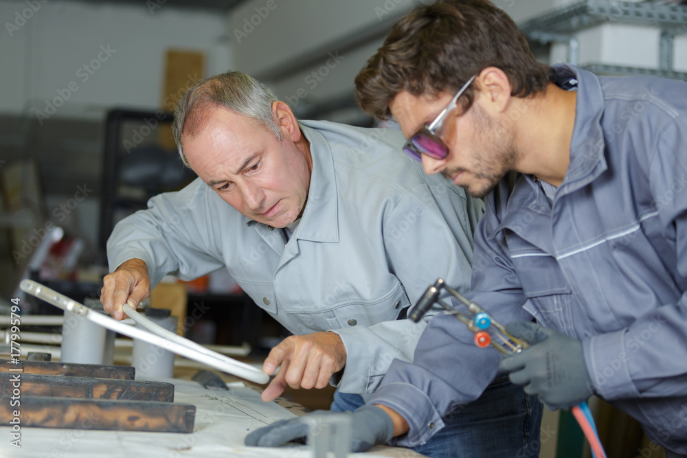 Two men in gray work uniforms are immersed in a welding training session at a workshop table. One uses a measuring tool, while the other holds a welding torch. Shelves and equipment are visible in the background, reflecting their dedication to precision and skill enhancement.