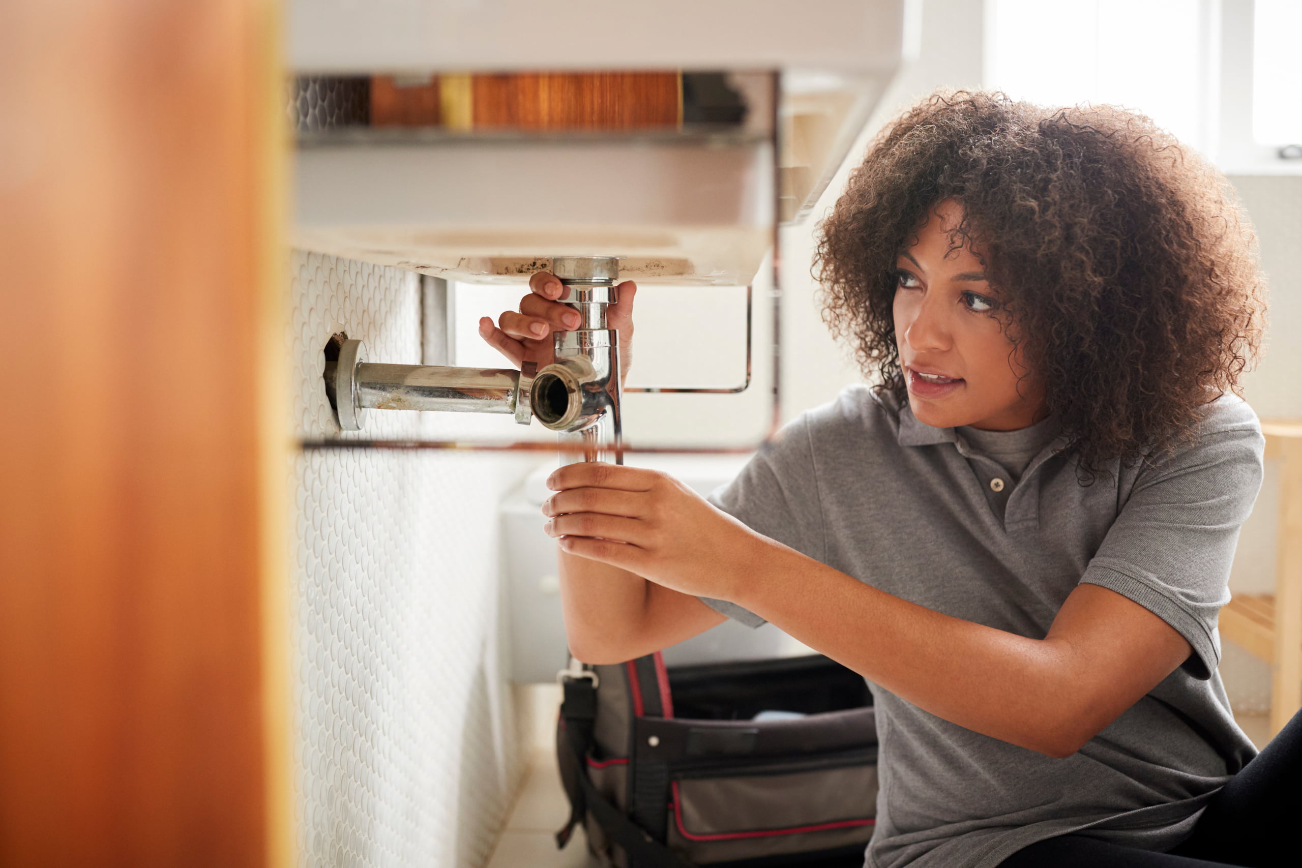 A young female plumber with curly hair sits on the floor, inspecting plumbing under a sink and holding a pipe wrench. Tools and a bag are scattered nearby, showcasing her dedication to getting the job done right.